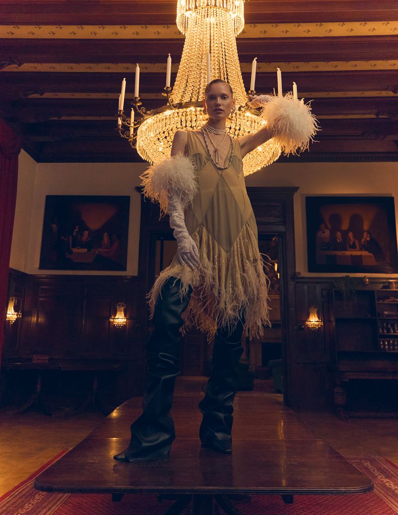 A fashion shot showcases a model wearing a feathered dress and thigh-high boots standing on a wooden table under a chandelier in a room.