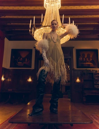 A fashion shot showcases a model wearing a feathered dress and thigh-high boots standing on a wooden table under a chandelier in a room.