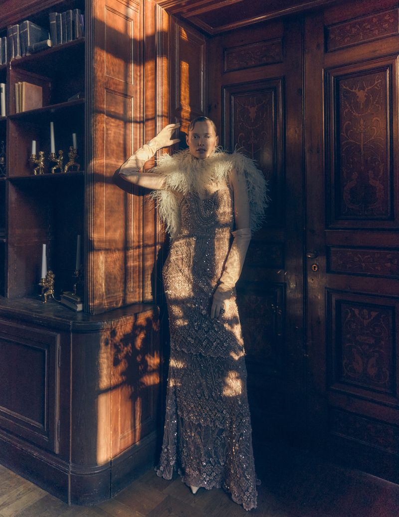 Elegant woman in a feather-trimmed gown poses indoors by a wooden door and bookcase in soft lighting, creating a luxurious ambiance.