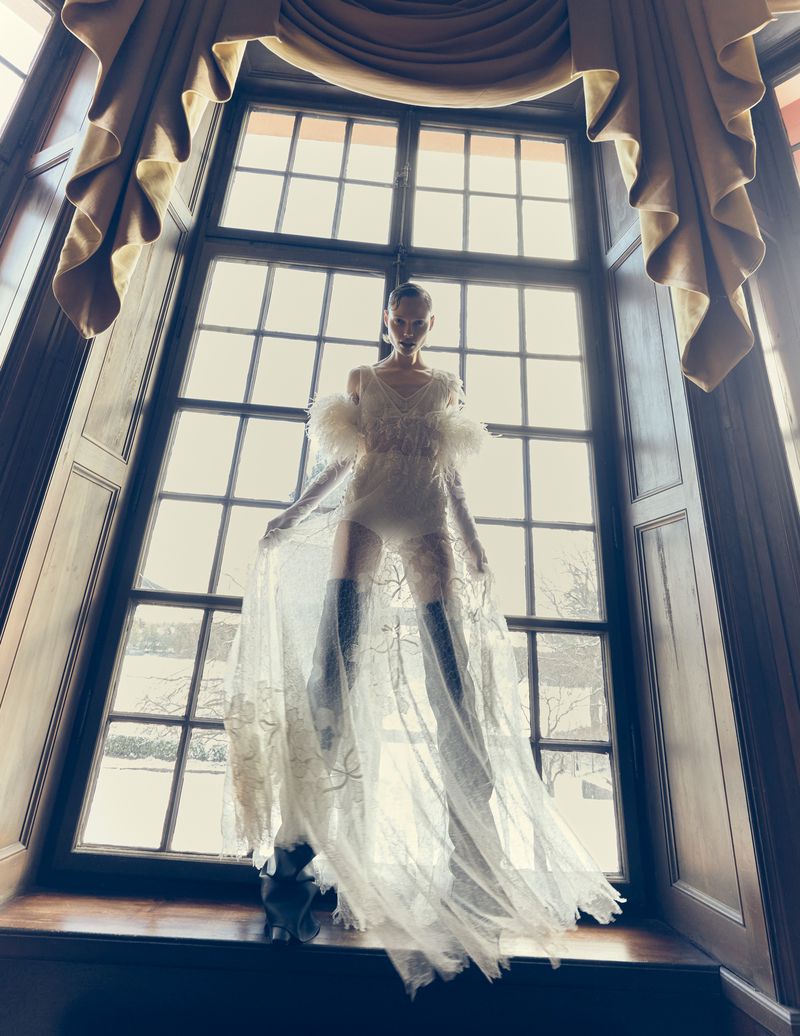 A slender woman stands in front of a large window with a sheer gown and thigh-high boots, bathed in soft natural light.
