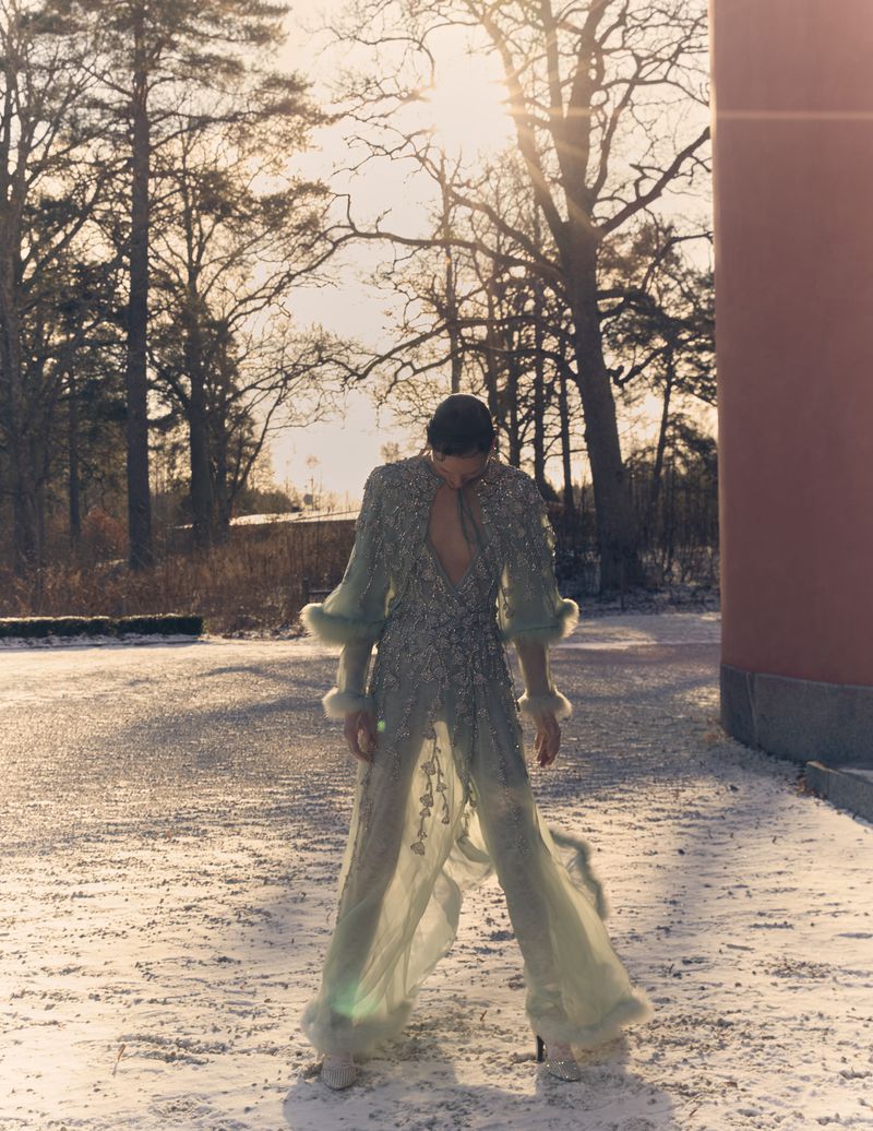 Woman in an embellished sheer gown with fur trim poses on a snow-covered path in a sunlit outdoor setting with bare trees and a column.