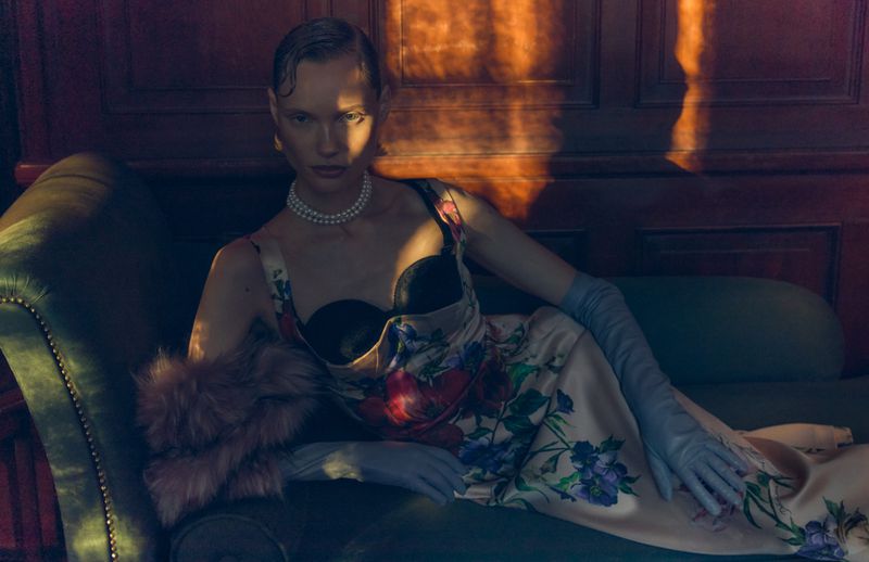 Woman in a floral dress with black corset and long gloves lounges on a green couch with soft lighting in an indoor setting.
