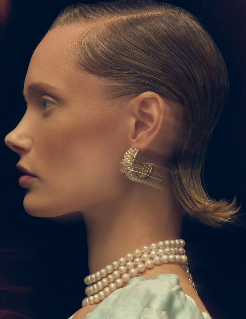 Close-up profile of an adult woman with combed-back light brown hair, wearing pearl earrings and necklace against a dark backdrop.