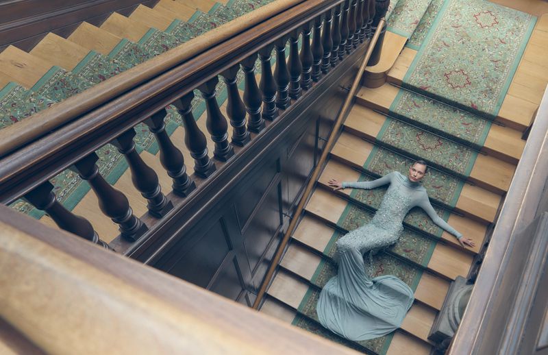 Woman in a flowing gown lying on a carpeted staircase. Indoor fashion shot with architectural details, brown wood, and soft, diffused light.
