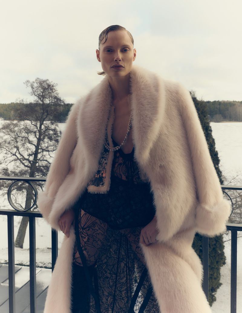 A blonde fashion model poses in a long, light pink fur coat over a black lace dress on a balcony with a snowy, tree-lined background.