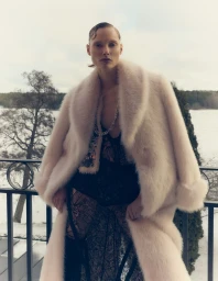 Model wearing a pale fluffy coat and dark lace outfit standing on a balcony overlooking a snowy lakeside landscape.