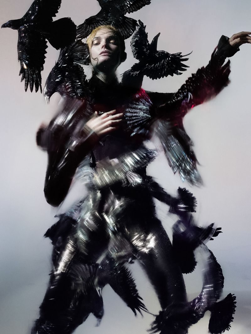 Fashion portrait of a blonde woman surrounded by black crows, wearing a shiny black, textured outfit against a pale gradient background.