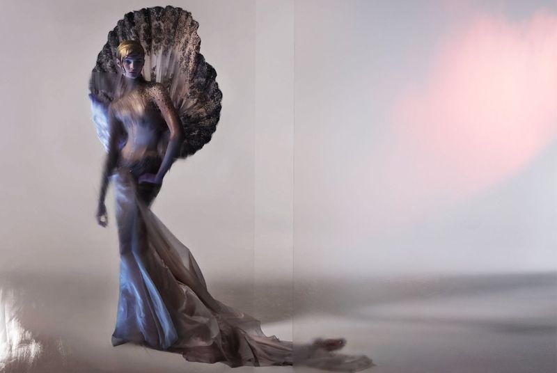 Woman in couture gown poses dramatically in a studio setting with feathered fan accentuating the flowing silhouette, creating a surreal, elegant image.