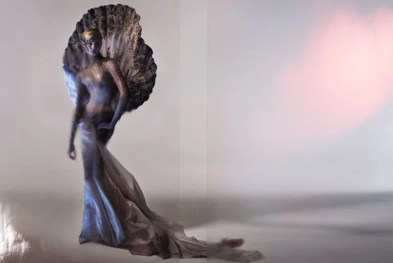 Woman in couture gown poses dramatically in a studio setting with feathered fan accentuating the flowing silhouette, creating a surreal, elegant image.