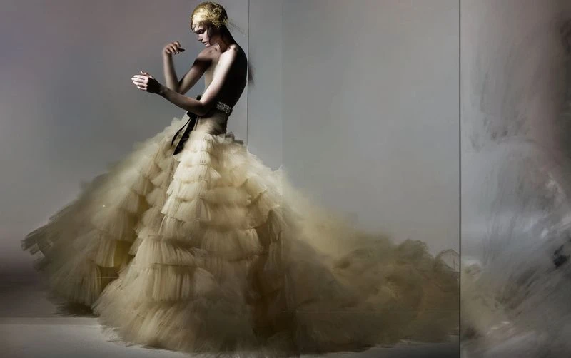 Elegant woman in a cream-colored layered gown, highlighted by a black sash, posing in a fashion studio against a light gray backdrop.