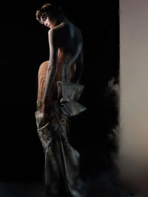 Adult female fashion model in an elaborate gown, shot in a dark photo studio. The model is looking down, with a large bow at her waist.