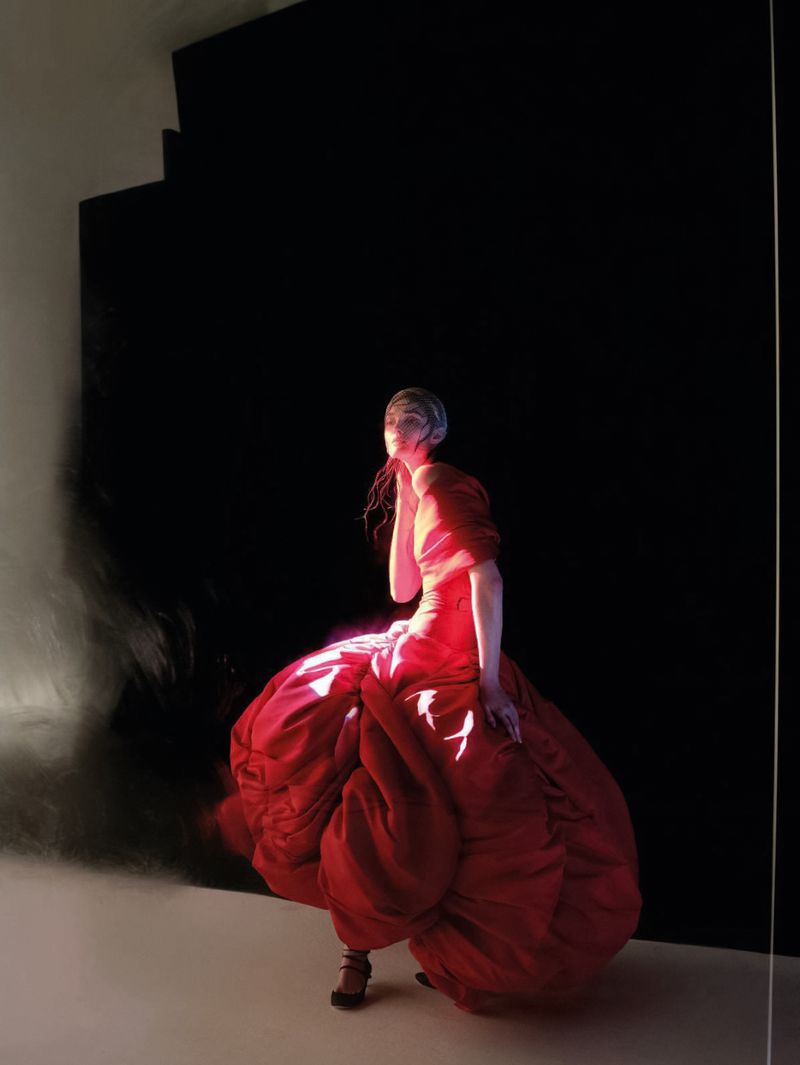 Woman in a red gown poses in a photo studio with black and white walls and floor, casting dramatic shadows from overhead lighting.