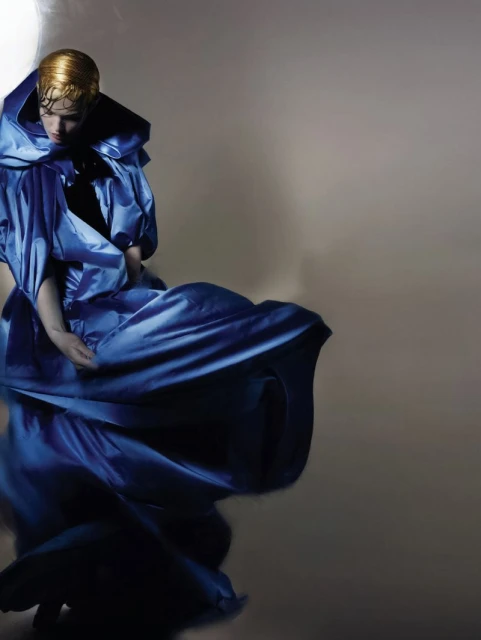 Fashion shot of a blond adult in a blue voluminous dress, kneeling in a photo studio with cool lighting, emphasizing the flowing fabric.