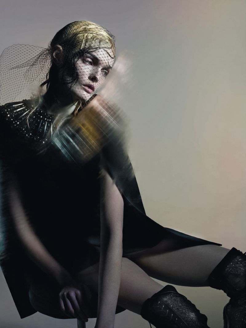 Fashion shot of a blonde woman in a dark dress and studded boots, seated and posing against a grey background with motion blur.