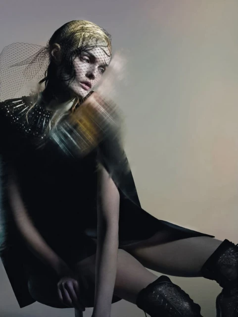 Fashion shot of a blonde woman in a dark dress and studded boots, seated and posing against a grey background with motion blur.