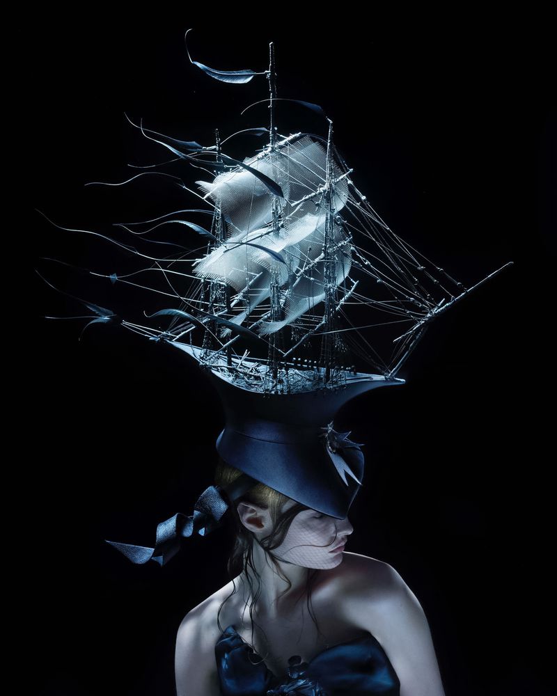 Woman in conceptual fashion portrait wearing a ship hat, looking down in dark studio. Fine art image with unique headwear and garment.
