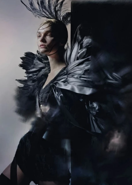 Dramatic fashion portrait of a model with feathered headwear and shoulder embellishments against a stark background, studio shot.