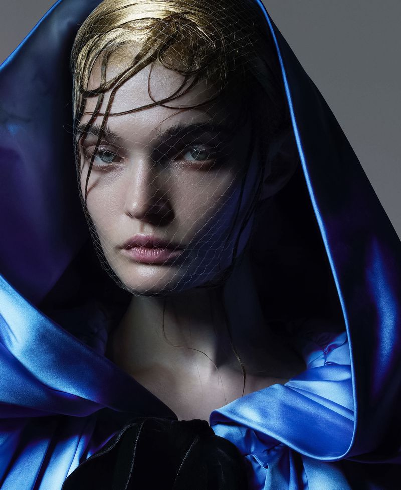 Close-up of a fair-skinned adult woman with blonde, slicked-back hair under a mesh veil, posing in an elegant blue satin hooded gown.