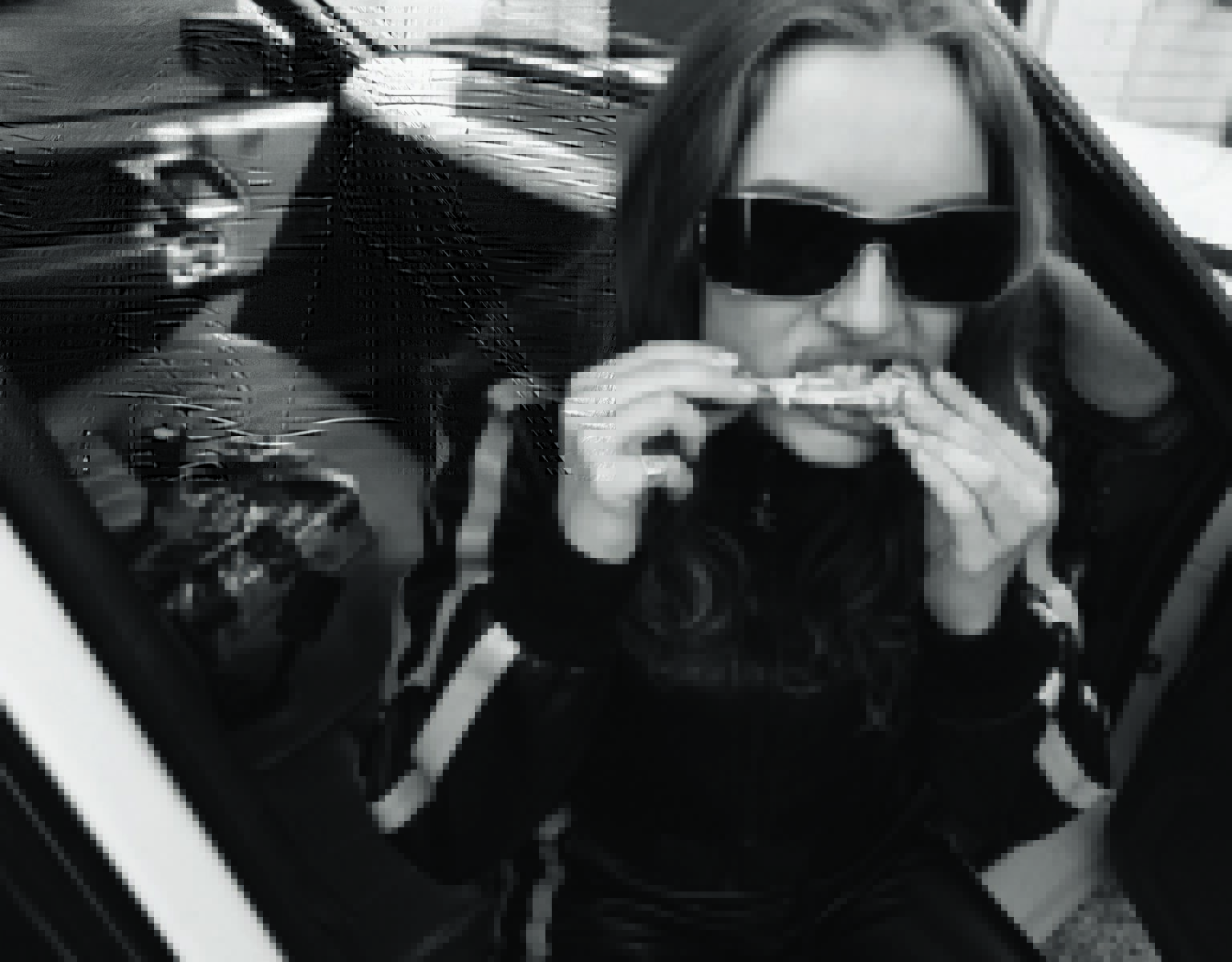 A young woman in sunglasses eats chicken wings inside a car in a black and white photograph.