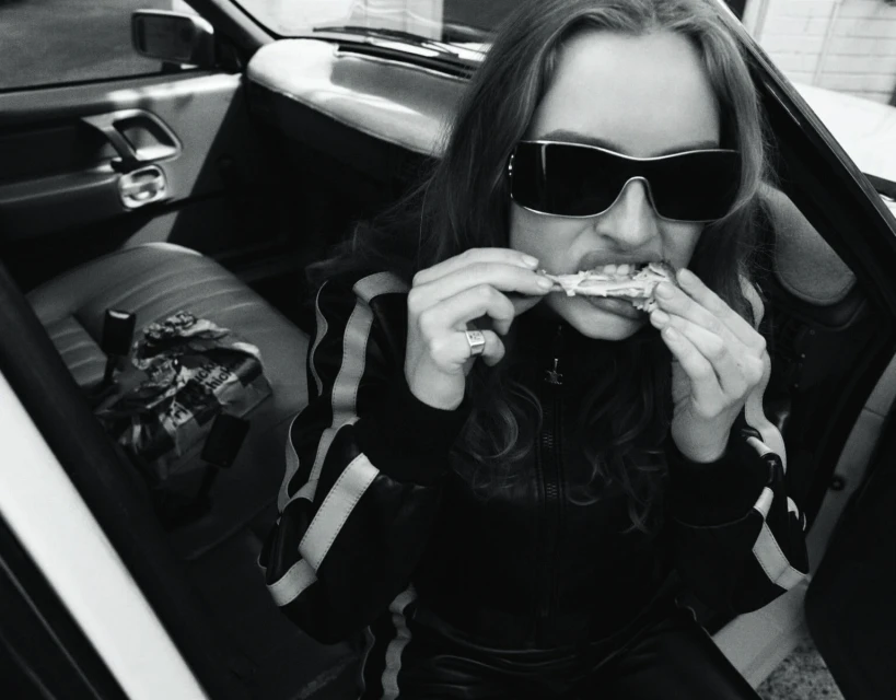 A young woman in sunglasses eats chicken wings inside a car in a black and white photograph.