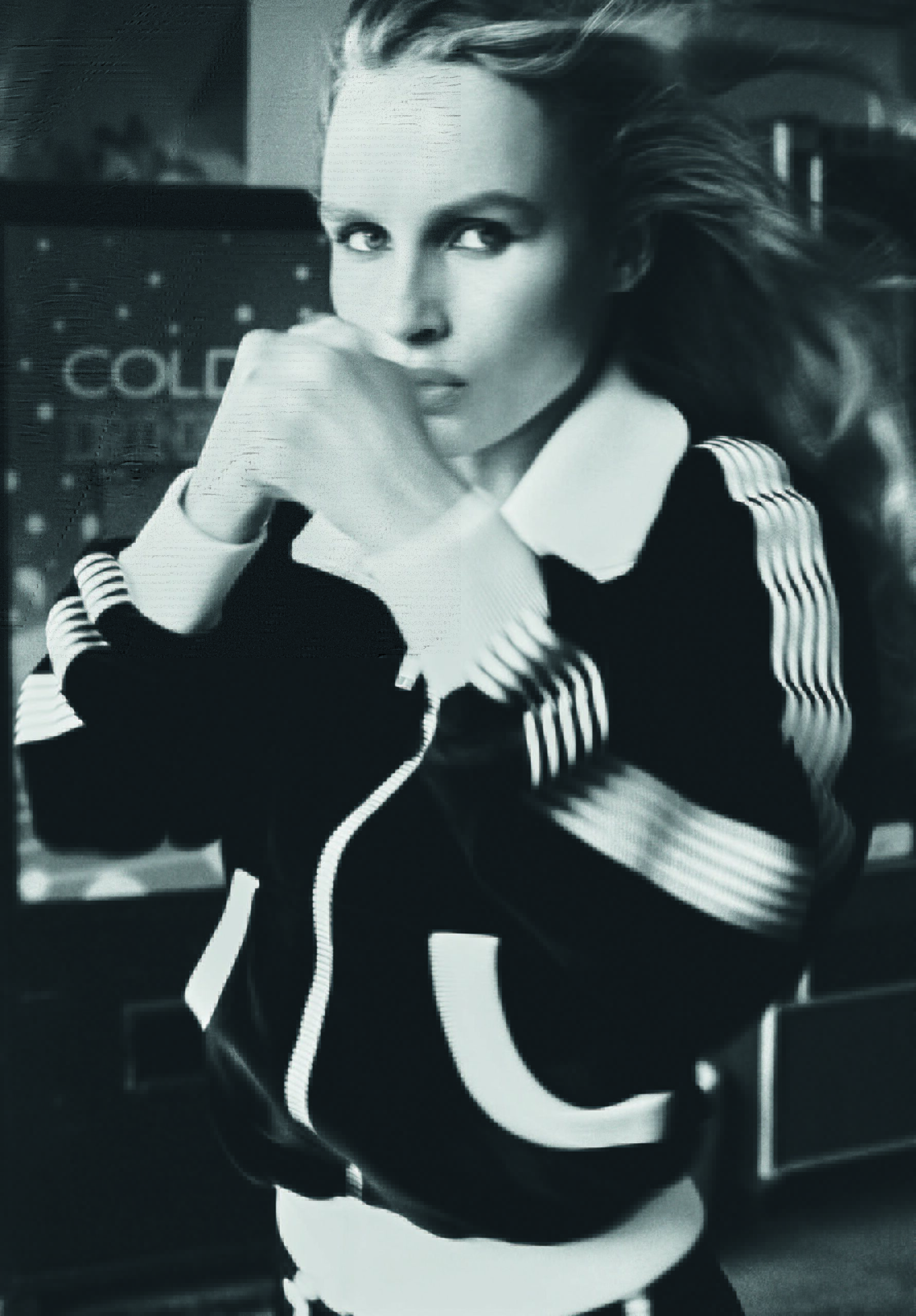 Black and white portrait of a blonde woman in an athletic jacket with fists up, posed in front of a soda machine indoors.