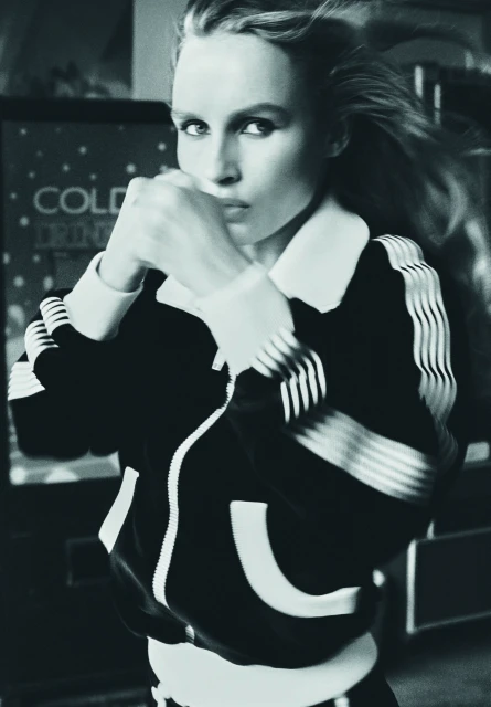 Black and white portrait of a blonde woman in an athletic jacket with fists up, posed in front of a soda machine indoors.