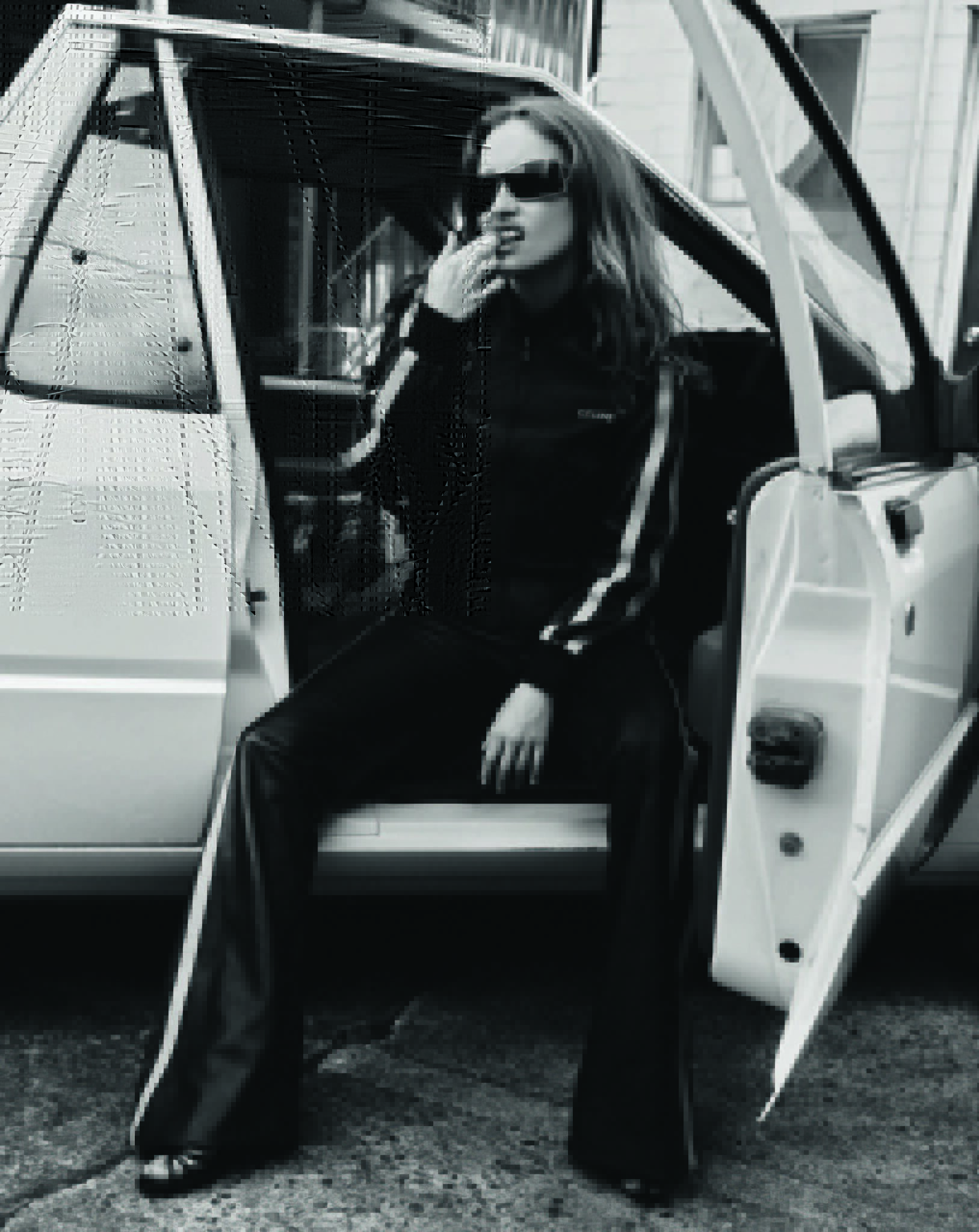 Black and white shot of a woman in sunglasses and sweatsuit sitting in an open car doorway, leaning back against the vehicle.