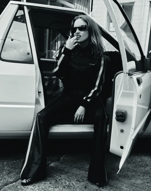 Black and white shot of a woman in sunglasses and sweatsuit sitting in an open car doorway, leaning back against the vehicle.