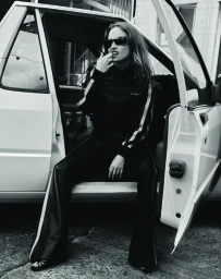 Woman in dark sunglasses and track jacket seated inside open car door