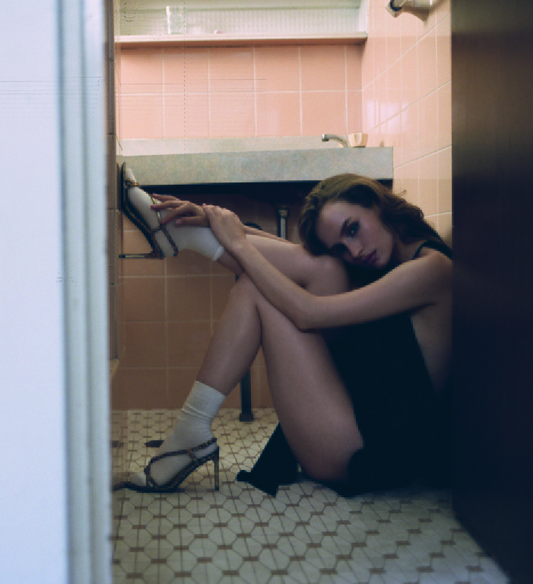 A young woman poses in a dark dress and high heels while seated on a patterned tile floor next to a pink-tiled bathroom wall and white door.