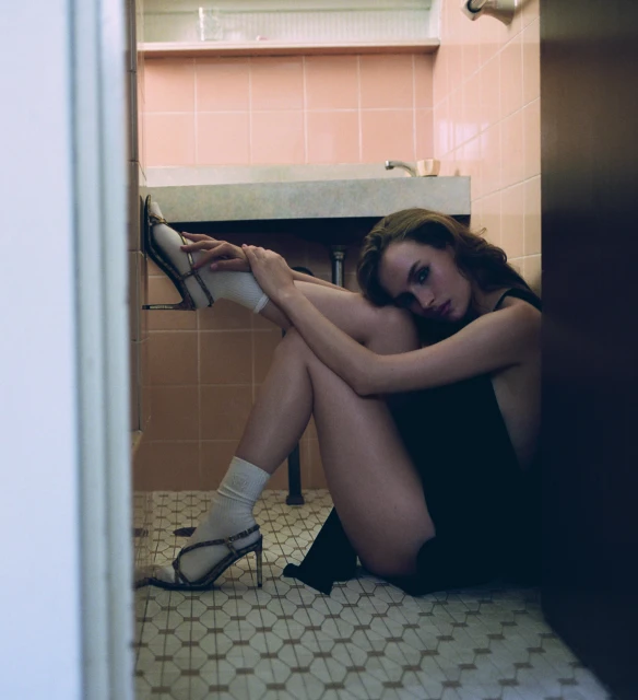 A young woman poses in a dark dress and high heels while seated on a patterned tile floor next to a pink-tiled bathroom wall and white door.