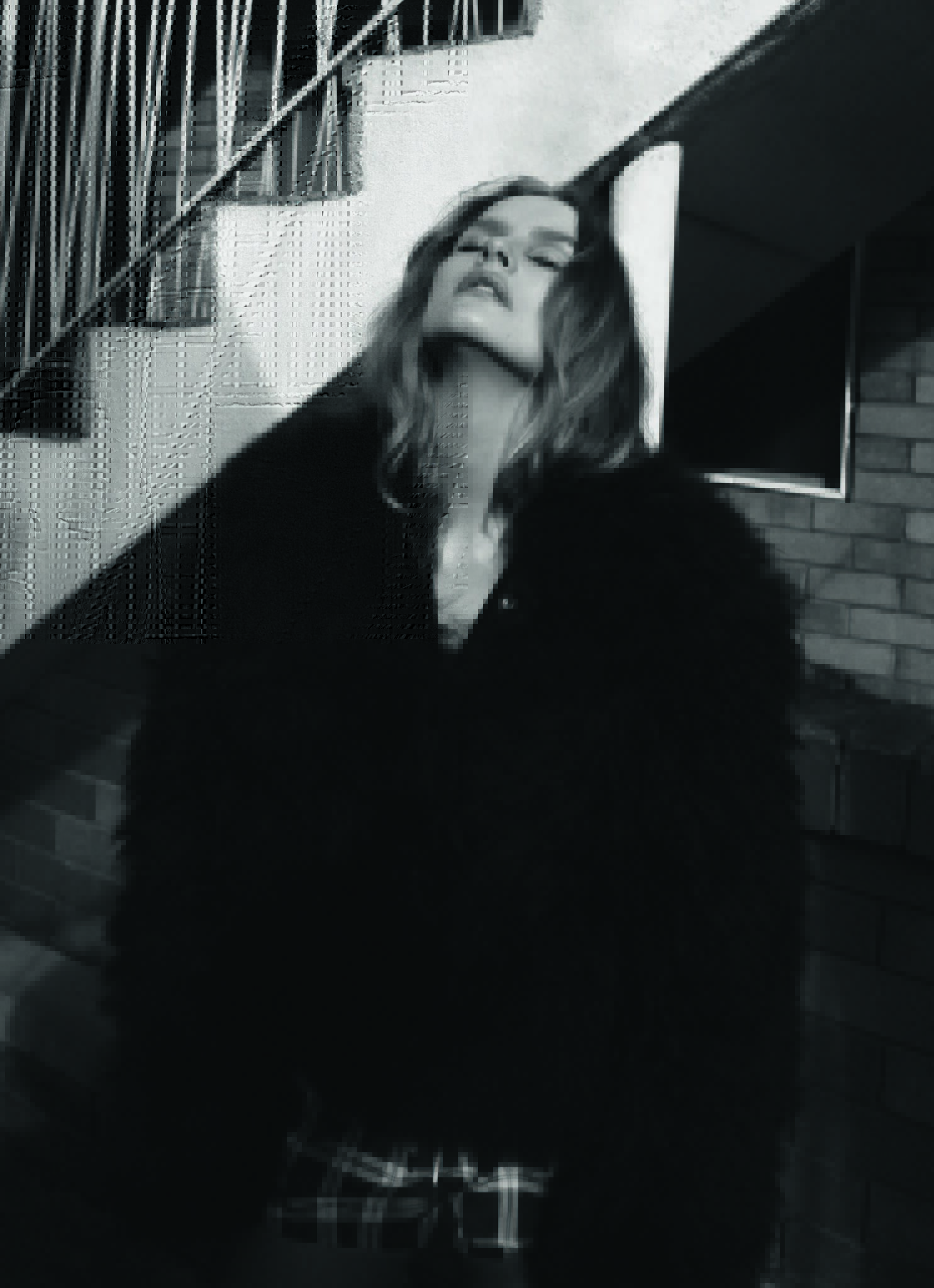 Black and white editorial portrait of a woman wearing a dark textured coat leaning against concrete stairs.