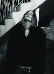 Black and white editorial portrait of a woman wearing a dark textured coat leaning against concrete stairs.