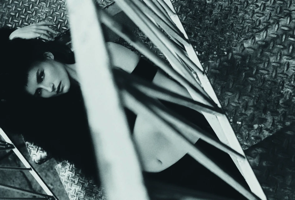 Black and white fashion portrait of a woman lying on a diamond plate surface behind metal bars, showcasing a raw and industrial aesthetic.