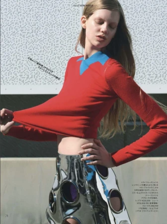 A fair-skinned model showcases a red sweater with a blue collar and a silver skirt with cutouts against a textured wall outdoors.