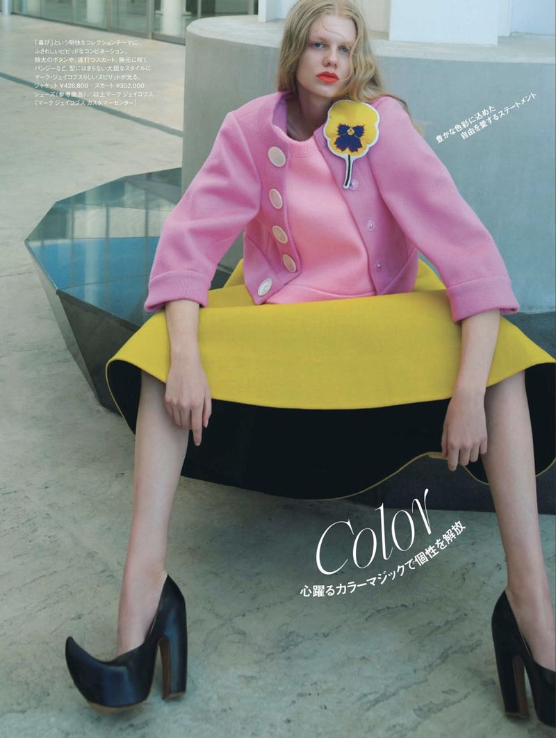 Woman in pink jacket and yellow skirt seated on a geometric bench in an urban setting, wearing unique black platform shoes. Fashion shot.