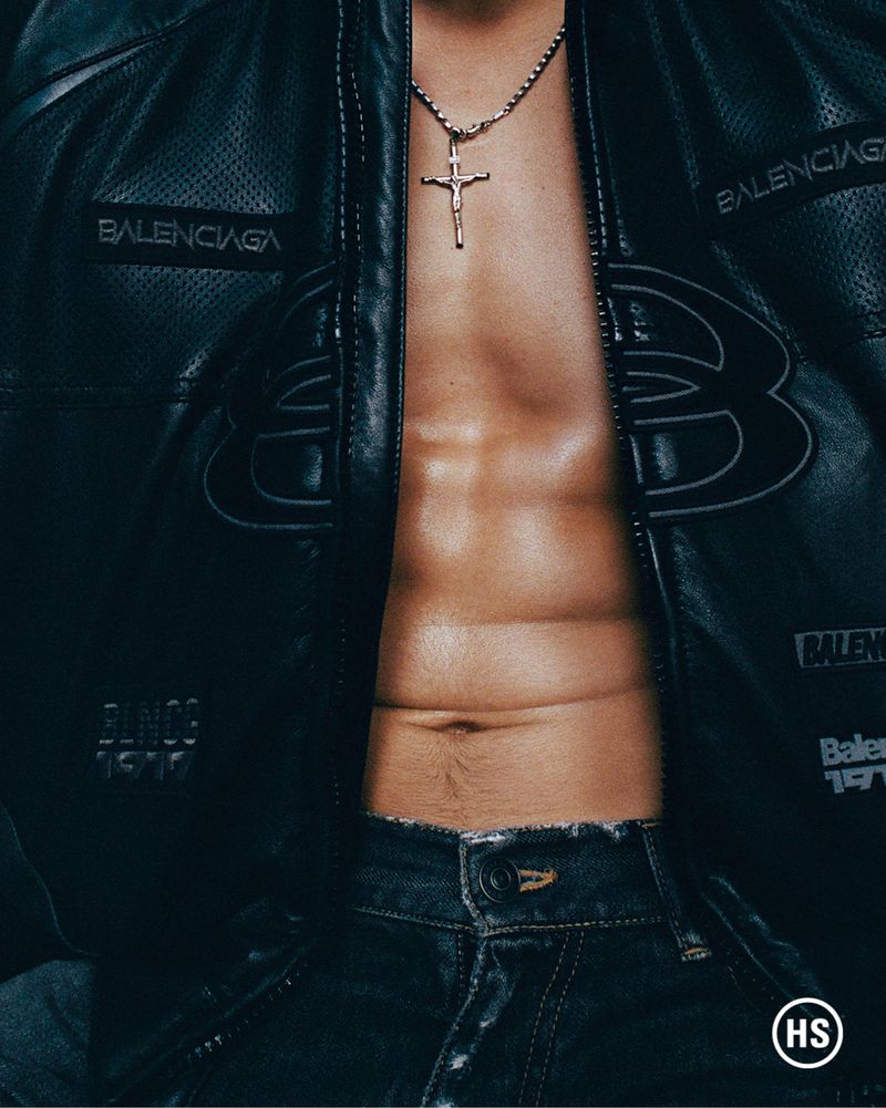 Close-up of a male torso wearing a black leather jacket over bare skin with a silver crucifix necklace and dark jeans.