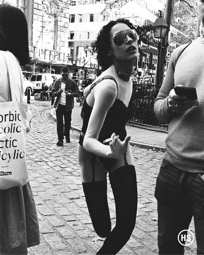 Black and white street-style photograph of a woman in lingerie and thigh-high stockings smoking a cigarette in an urban setting.