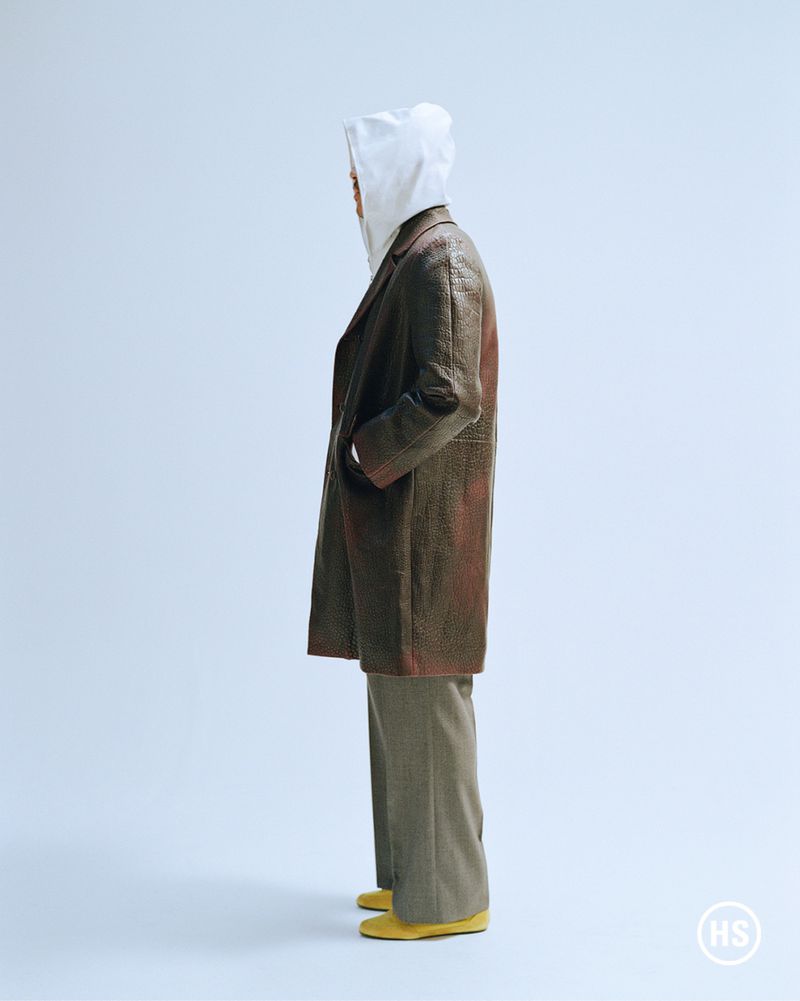 Fashion shot featuring a slender adult wearing a hooded shirt, coat, pants, and shoes. The model faces to the side in a photo studio.