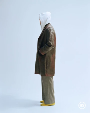 Fashion shot featuring a slender adult wearing a hooded shirt, coat, pants, and shoes. The model faces to the side in a photo studio.