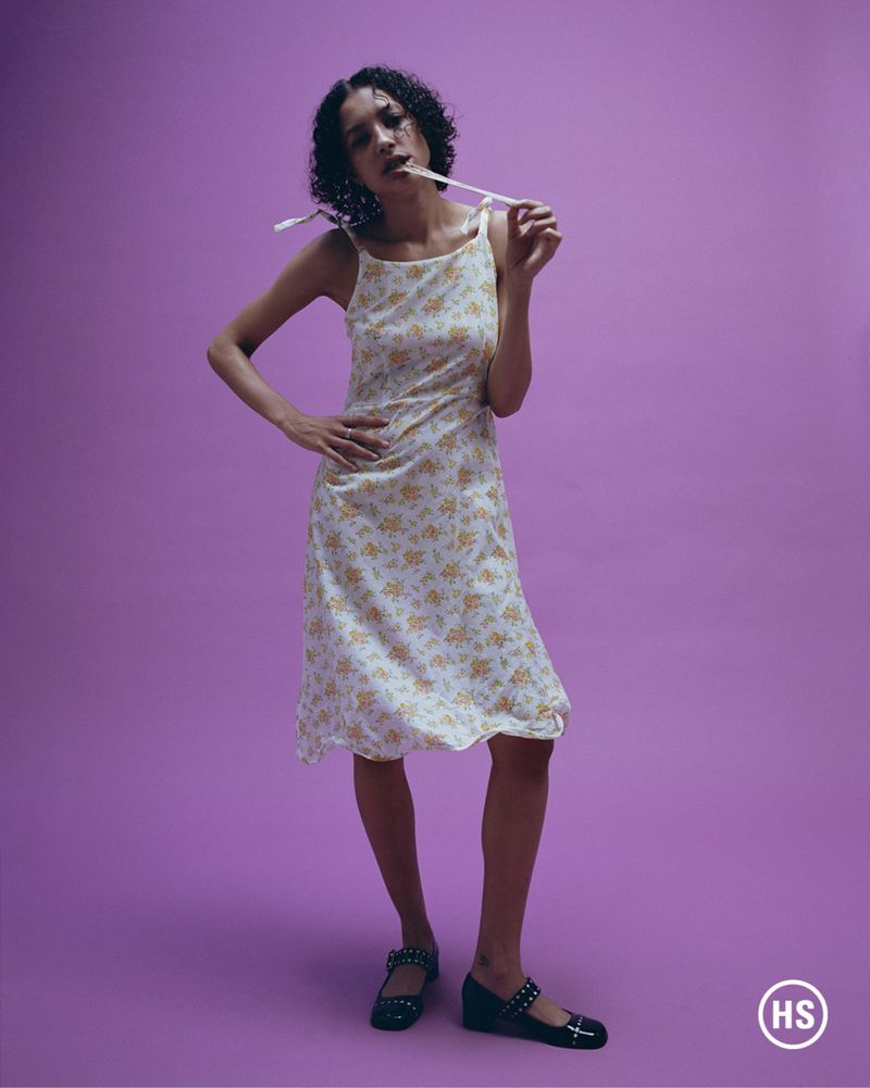 A young woman in a floral dress poses on a purple background, holding a white stick in her mouth in a studio shot.