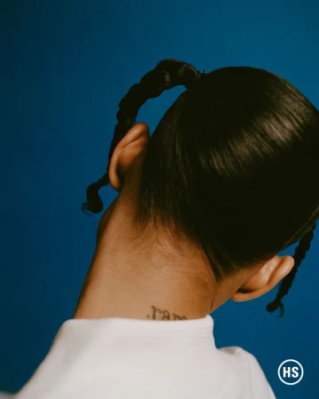 Close up studio shot of the back of a woman's head with braids and a neck tattoo against a solid blue background. High-quality lighting.