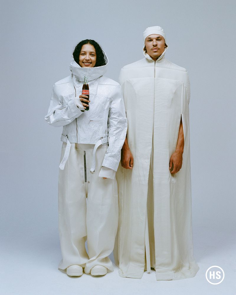 Two people in white textured clothing pose indoors in a studio. One person holds a Coca-Cola bottle. Fashion and beverage advertisement.