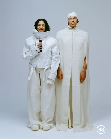 Two people in white textured clothing pose indoors in a studio. One person holds a Coca-Cola bottle. Fashion and beverage advertisement.