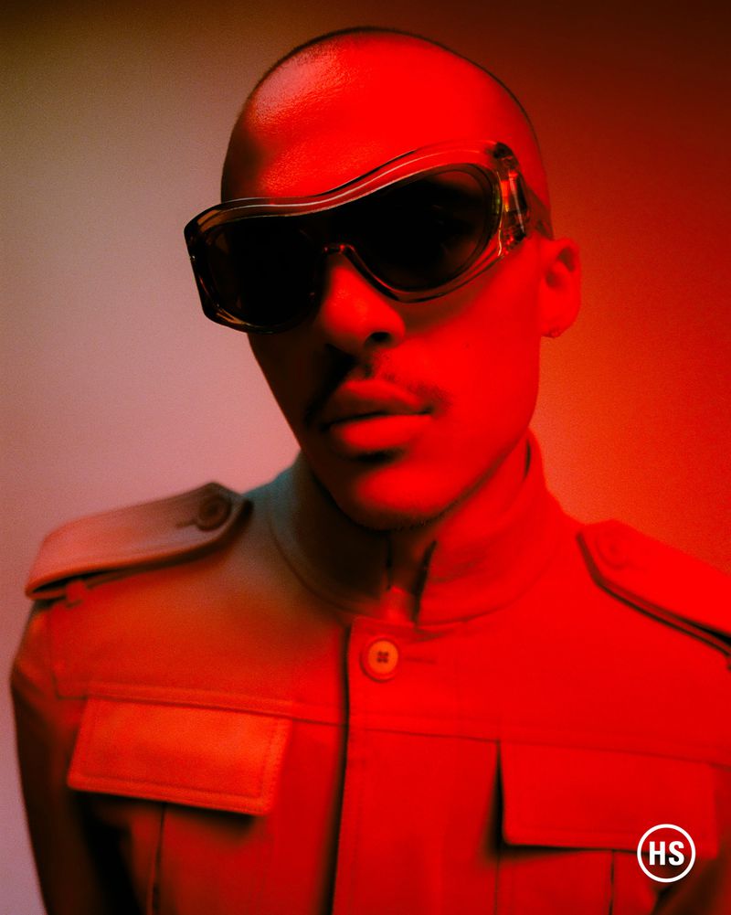 Close-up portrait of a model wearing dark futuristic sunglasses and a tan jacket under vibrant red studio lighting.