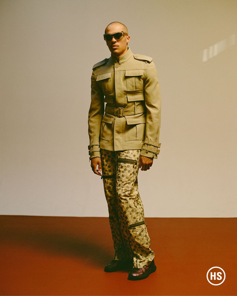 Man in khaki jacket and patterned pants standing in a studio with warm lighting and sunglasses, fashion portrait.