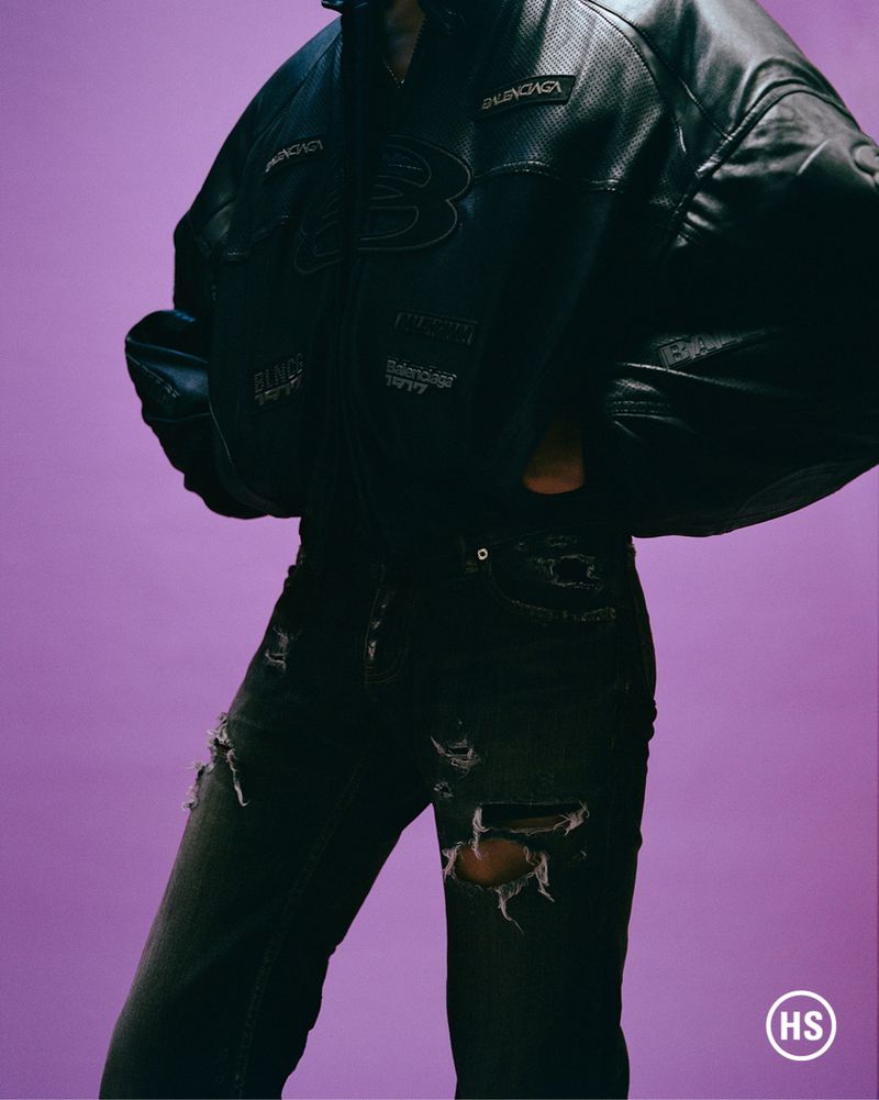 Person in black leather Balenciaga jacket and ripped jeans posing against a purple background in a fashion studio shot.