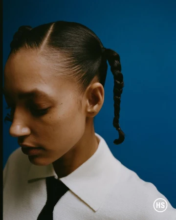 Close-up portrait of a model with braided pigtails wearing a white collared shirt and black tie against a blue backdrop.