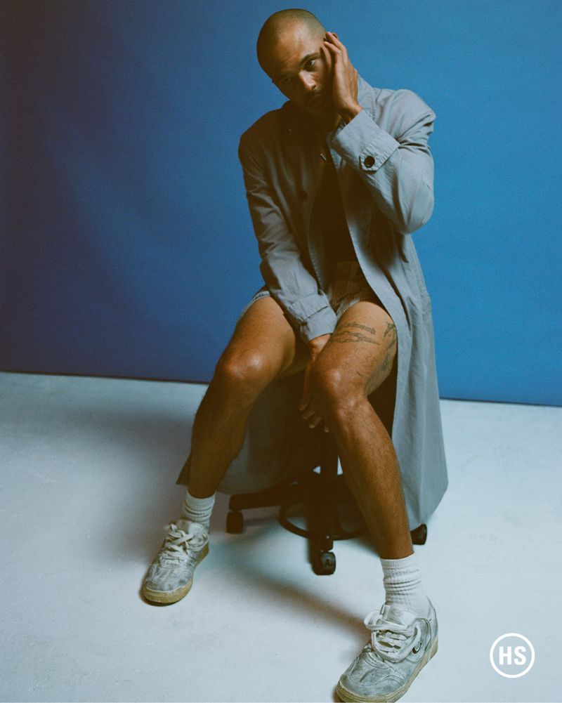 Bald adult male in a gray jacket poses seated on a chair in front of a blue background, wearing white socks and shoes.