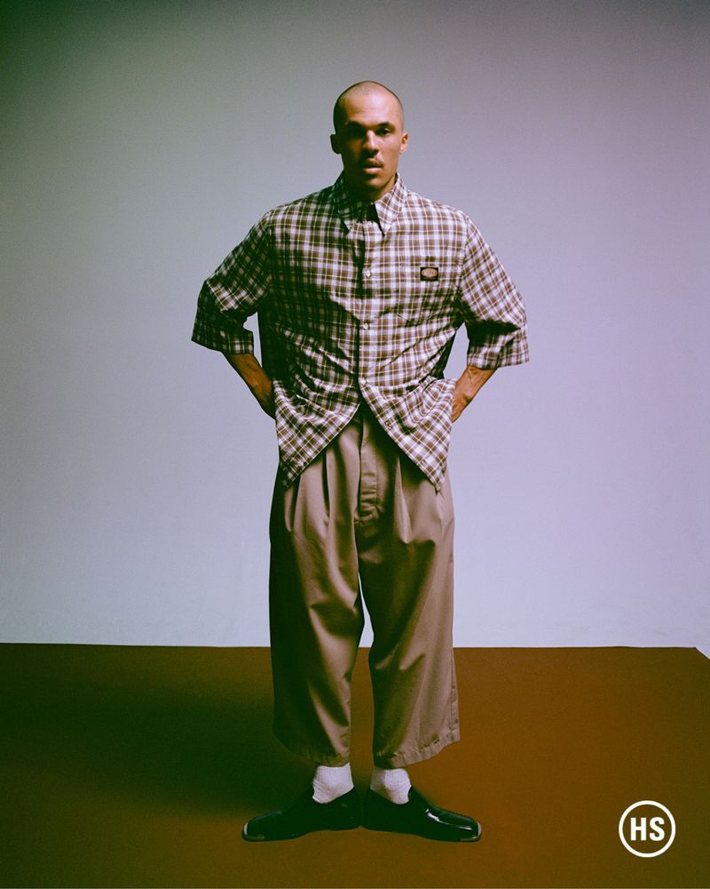Man in oversized shirt and pants poses in studio with hands on hips, standing on brown floor against pale blue background, fashion shoot.